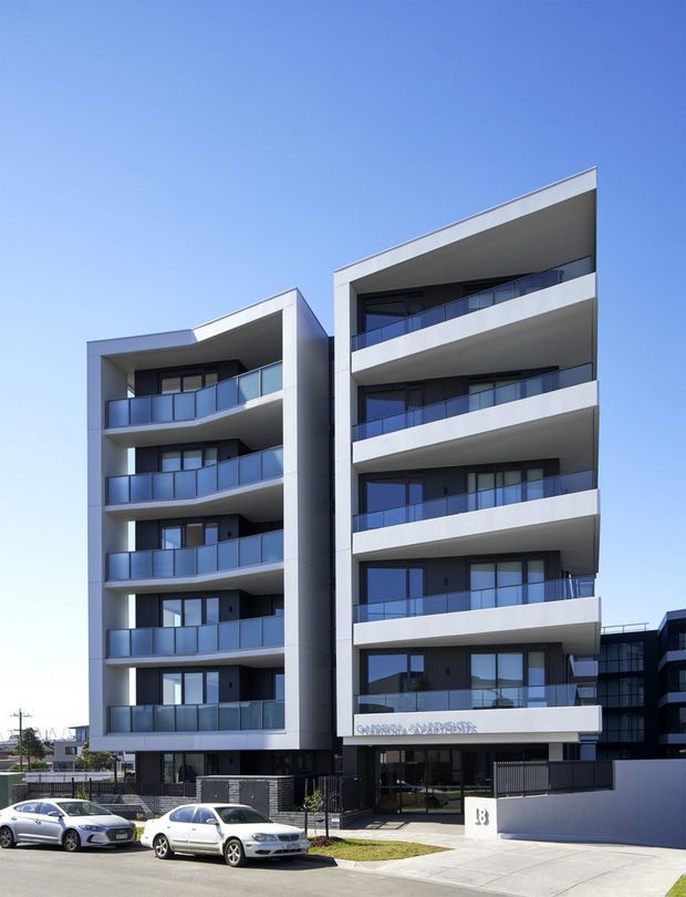 Street-level exterior photograph of a contemporary multi-storey apartment building with two articulated wings, white rendered or panelled facade surfaces, dark recessed walls, extensive glazed balconies with blue-grey balustrades, and a flat roofline. The building entrance and street frontage are visible, with neighbouring urban development and parked cars in the foreground.