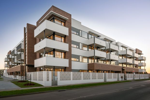 Exterior architectural photograph of a contemporary multi-storey apartment building viewed from the street. The development features white rendered and panelled facades, prominent projecting balconies with dark metal balustrades, extensive glazing, and contrasting reddish-brown brick vertical sections and parapets. A decorative perforated masonry boundary wall and landscaped verge sit along the street frontage.