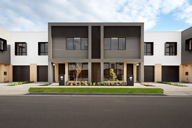 Front exterior view of a contemporary multi-unit residential townhouse development. The symmetrical facade combines crisp white rendered walls, dark garage doors and window frames, charcoal horizontal louvres, pale masonry accents, and recessed entry patios. A paved forecourt with narrow planted strips fronts the building, with a broad asphalt street in the foreground.