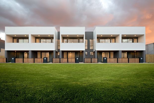 Front exterior view of a row of attached contemporary multi-storey townhouses, featuring repeated white box-like upper volumes, dark grey lower sections, recessed balconies or terraces, large horizontal windows, timber-look privacy fencing, and small front entries. The development is set behind a broad lawn under a dramatic cloudy evening sky.