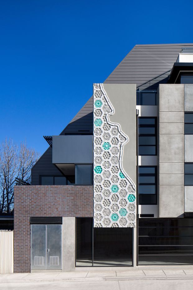 Street-level exterior photograph of a contemporary multi-storey building with a steep dark metal roof, projecting balconies, exposed concrete panels, dark glazing, and a prominent decorative facade screen featuring geometric hexagonal patterns with turquoise accents. A brick boundary wall and entry glazing sit at ground level.