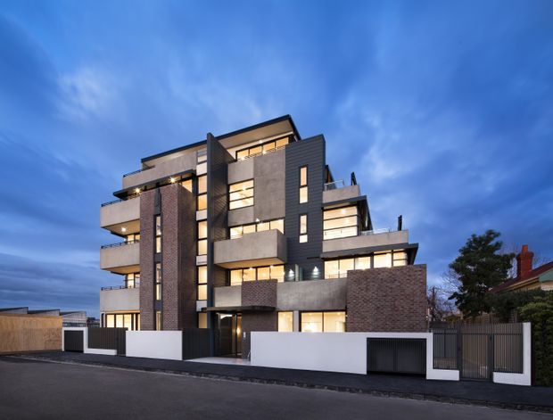 Exterior dusk photograph of a contemporary multi-storey residential apartment building. The facade combines dark vertical cladding, exposed brick, pale rendered or concrete panels, extensive illuminated glazing, projecting balconies, and a rooftop terrace-like form. A secure street-level boundary wall and garage entrance occupy the foreground, with a neighbouring house visible at the side.
