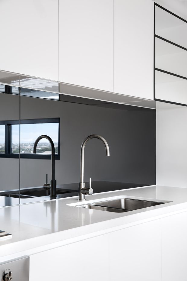 A close-up view of a sleek contemporary kitchen featuring a white island with an integrated stainless-steel double sink and two curved gooseneck mixer taps. Glossy white cabinetry extends to the ceiling, while a dark charcoal backsplash and black-framed window create strong contrast. A large mirror reflects the sink and cabinetry, and a stainless-steel appliance is partially visible below the benchtop.