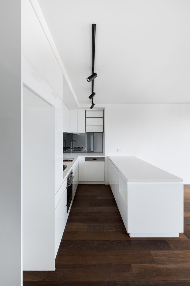 A bright minimalist white kitchen with an L-shaped run of handleless cabinetry and a large white island with an integrated cooktop or work surface. Dark timber flooring provides contrast, while a black linear track light with adjustable cylindrical spotlights hangs above. The space has clean planar surfaces, a concealed or integrated rangehood area, and a restrained monochrome palette.