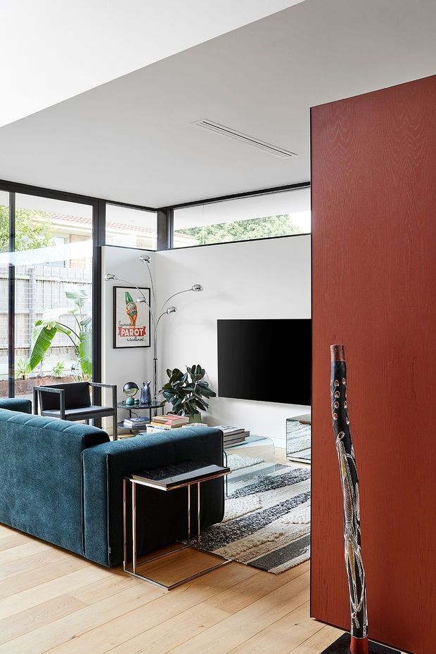 A bright, contemporary living room viewed from an adjacent space, featuring deep blue upholstered seating, a wall-mounted television, a black-framed glazed corner with garden views, pale timber flooring, a layered patterned rug, and a large reddish timber or wood-finish partition panel in the foreground. A slim linear air-conditioning vent and sculptural floor lamp add to the clean-lined interior.