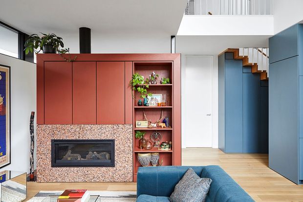 Bright contemporary living room with a blue sofa in the foreground, a built-in fireplace framed by a terrazzo-like feature wall, and terracotta-red cabinetry with open display shelving. Light timber flooring leads to a stairway and blue built-in joinery at the rear, with a mezzanine or upper-level void visible above.