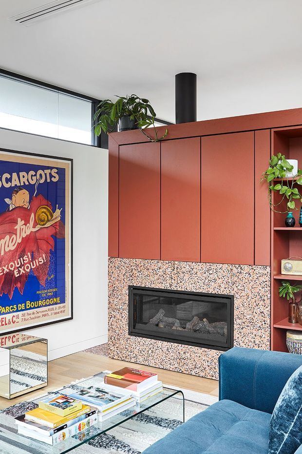 Contemporary living room featuring a built-in terracotta-red fireplace wall with integrated cabinetry and open shelving, a speckled terrazzo fireplace surround, black firebox and flue, large windows, a blue upholstered sofa, glass coffee table, patterned rug, plants, and framed graphic artwork.