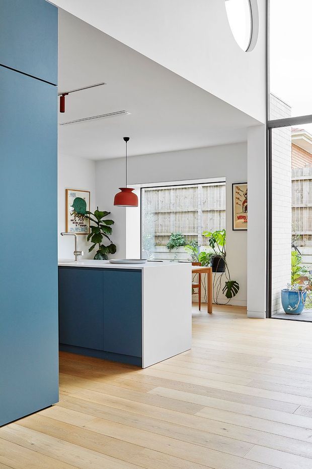 Bright contemporary open-plan kitchen interior with a compact blue island, white cabinetry, pale timber flooring, and a red pendant light. Full-height glazing opens toward a planted courtyard or lightwell, while artwork and indoor plants add colour. The room has a clean, airy appearance with a partially visible high ceiling and strong indoor–outdoor connection.