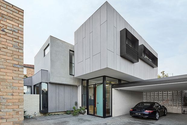 Exterior photograph of a contemporary multi-level residence with a sharply pitched, folded roof form and projecting upper floor clad in pale grey vertical panels. The home combines exposed brickwork, smooth grey rendered walls, extensive dark-framed glazing, and projecting dark metal window boxes. A covered carport with perforated block screening occupies the foreground, with a paved driveway and a visible entry door beneath the cantilevered upper level.