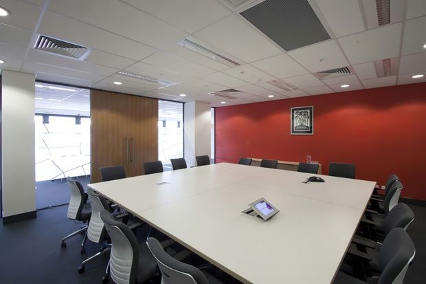 A modern commercial boardroom with a large white conference table surrounded by ergonomic office chairs. The room has a bold red feature wall with framed artwork, timber double doors, expansive glazed windows, dark carpet, and a suspended grid ceiling with recessed lighting and air-conditioning vents.