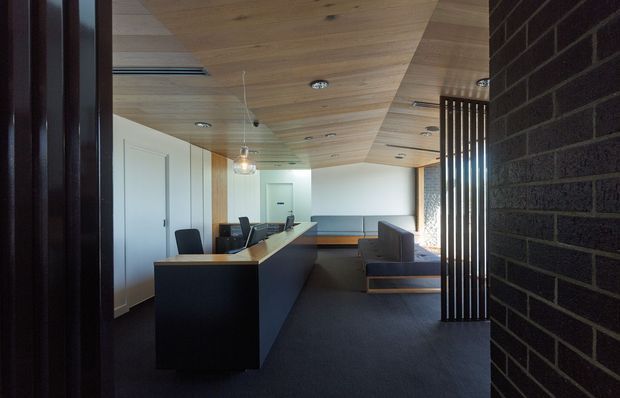 A refined contemporary open-plan interior viewed through a dark brick entry, featuring a long black kitchen island or built-in counter, dark carpeted flooring, a warm timber-lined ceiling, white walls and cabinetry, pendant and recessed lighting, and a dark-framed vertical slatted room divider. A compact seating area is visible beyond the counter.
