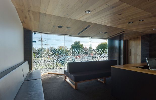 A contemporary office reception or lounge area with a dark carpeted floor, timber-lined ceiling, and large patterned glazed windows. A long light-grey sofa sits along the left wall, facing a dark charcoal sofa with timber legs. A dark reception counter or partition occupies the foreground, while a timber-lined doorway and dark vertical slatted feature wall define the rear of the space. The glazing provides strong daylight and views toward an urban streetscape.