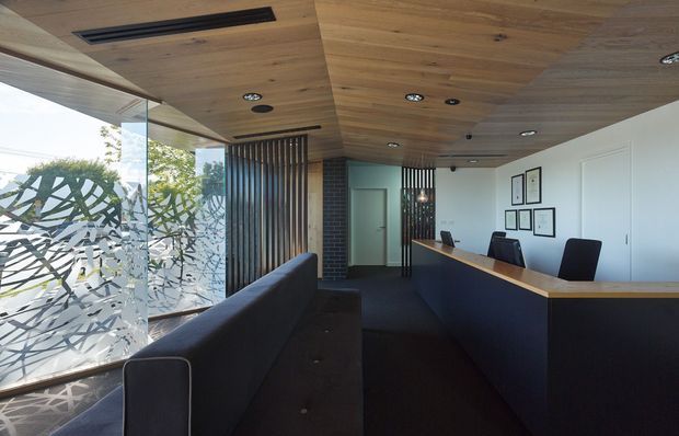 A contemporary reception or front-desk area with a long dark built-in counter and timber benchtop, upholstered dark seating, a timber-lined ceiling, white walls, and recessed lighting. Full-height glazing with patterned privacy film and vertical timber screening bring daylight into the space, while a dark brick column and framed artwork add contrast.