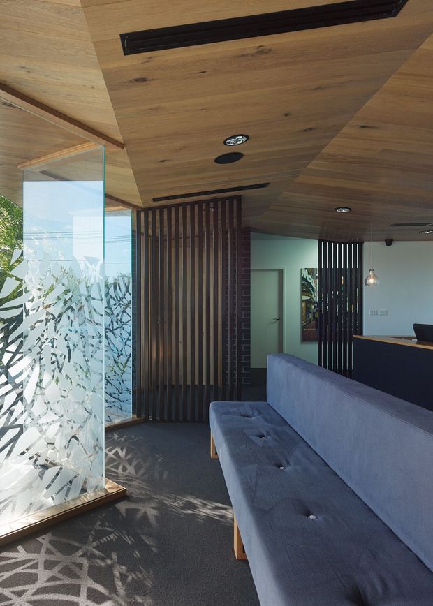 A contemporary interior reception or waiting area with a long blue-grey upholstered sofa, dark carpet, timber-lined ceiling, recessed lighting and linear air vents. Vertical dark timber battens create partial screens around a doorway and counter area, while a large patterned translucent glass panel filters strong daylight and casts geometric shadows across the floor.