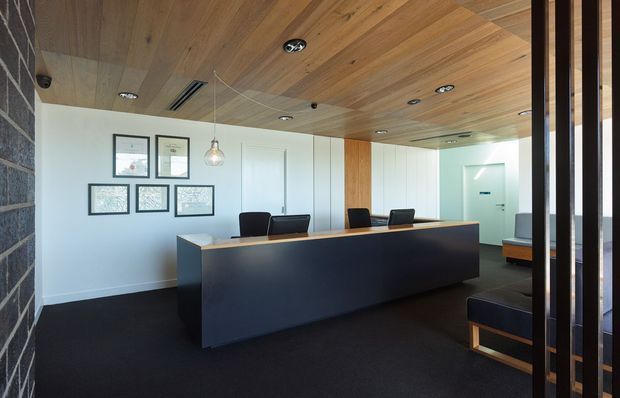 Contemporary reception or front desk area with a long dark built-in counter, black carpet, white walls, timber-lined ceiling, recessed downlights, framed artwork, pendant lighting, and dark stone-like wall cladding. Glazed and timber-framed partitions appear at the right, with seating visible behind the desk.