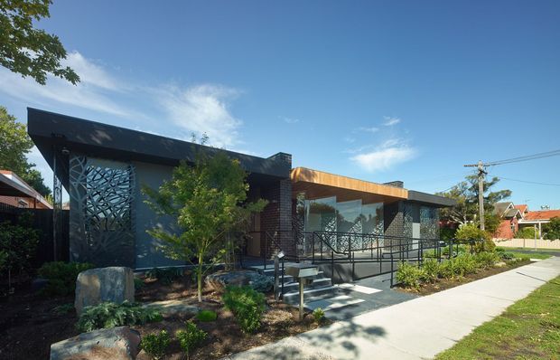 Single-storey contemporary building viewed from the street, with a low-slung flat roof, dark cladding and brickwork, a warm timber-toned fascia, extensive glazing, and a decorative patterned screen at the entrance. A broad accessible ramp with metal balustrades leads to the front door, surrounded by landscaped garden beds and a concrete footpath. Residential surroundings are visible in the background.