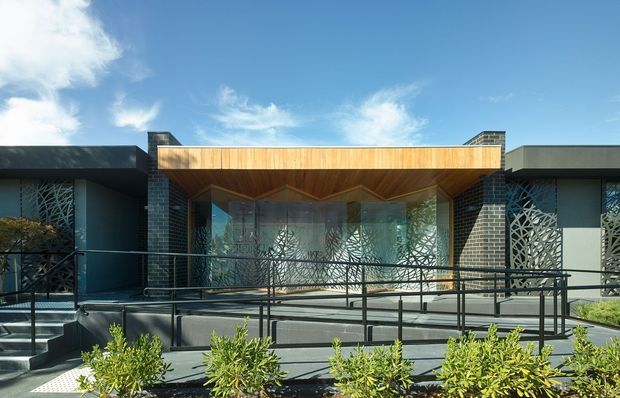 Front exterior view of a low-rise contemporary building with dark brick and rendered wall sections, a broad timber-lined projecting entrance canopy, patterned privacy screens, extensive glazing, and accessible ramped approaches with black metal balustrades. Low planting borders the paved forecourt beneath a bright sky.
