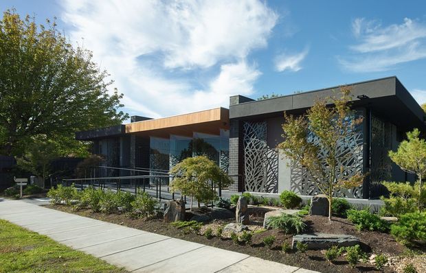 Contemporary single-storey building exterior with a broad flat roof, dark charcoal cladding, warm timber soffit or fascia, extensive glazing, and decorative laser-cut screens featuring organic branching patterns. The landscaped frontage includes raised planting beds, rocks, shrubs, and a concrete pedestrian path. A ramp and railings provide accessible entry.