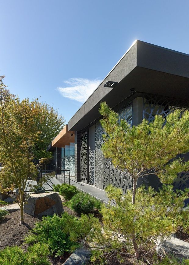 A contemporary single-storey building viewed from a landscaped approach. The facade features a deep dark flat roof canopy, textured dark cladding, extensive glazing, and distinctive decorative perforated metal screens with an organic branching pattern. A timber-toned projecting canopy marks the entrance, reached by a ramp and steps with black metal handrails. Native-style planting, rocks, and small trees surround the building under a clear blue sky.