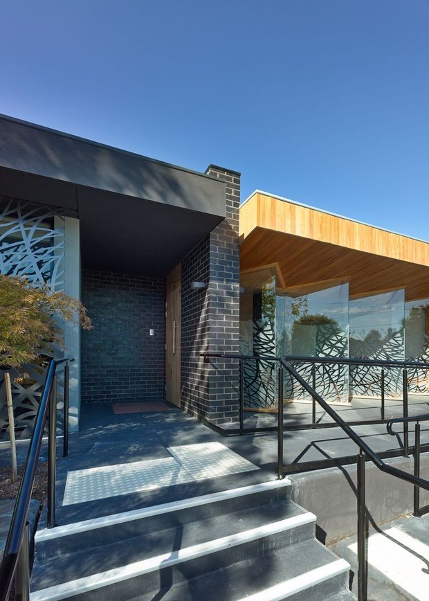 Exterior view of a contemporary building entrance approached by stairs and an accessible ramp. The facade combines dark brick masonry, charcoal metal soffits, warm timber cladding and a broad glazed frontage. Decorative patterned screens and black metal handrails define the entry, with strong sunlight and sharp shadows across the steps and landing.