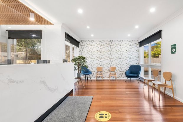 Bright contemporary reception or waiting area with polished timber flooring, white walls, a marble-look reception counter, patterned botanical feature wallpaper, blue lounge chairs, timber chairs, and abundant recessed lighting. A timber slatted ceiling feature and black roller blinds add contrast.