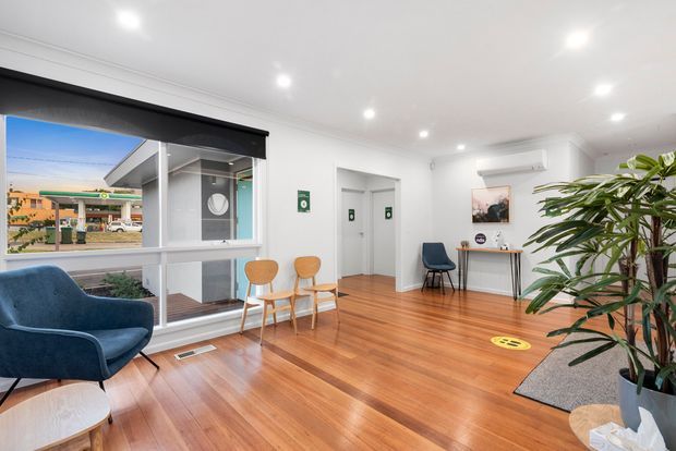 Bright, clean reception or waiting area with polished timber flooring, white walls and ceiling, large street-facing windows, a dark roller blind, blue armchairs, timber guest chairs, a narrow console table, indoor plants, and recessed ceiling lighting. The space appears to be a small commercial office or clinic entry area.