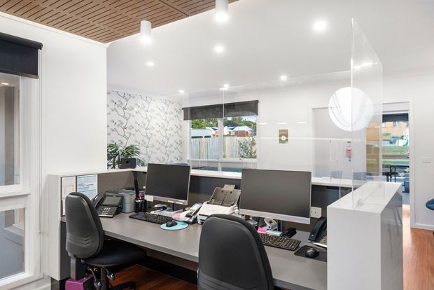 A bright, modern commercial office or reception workspace with multiple computer workstations arranged around a shared desk. The room has white walls, a patterned feature wall, timber slatted ceiling panels, recessed and pendant lighting, wood-look flooring, roller blinds, and a clear partition or screen near a white reception counter.