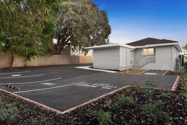 Exterior view of a small single-storey grey building beside a marked asphalt parking area, with a low-pitched dark roof, horizontal cladding, covered entry steps, accessible parking markings, timber fencing, and landscaped planting.