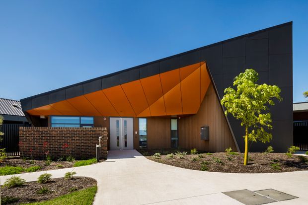 Modern single-storey building facade viewed from the street, featuring a dramatic angular projecting canopy with a vivid orange soffit, dark charcoal cladding, warm vertical timber-look wall cladding, and glazed entry doors. A low dark brick wall, landscaped garden beds, young tree, and concrete pedestrian path frame the entrance under a clear blue sky.