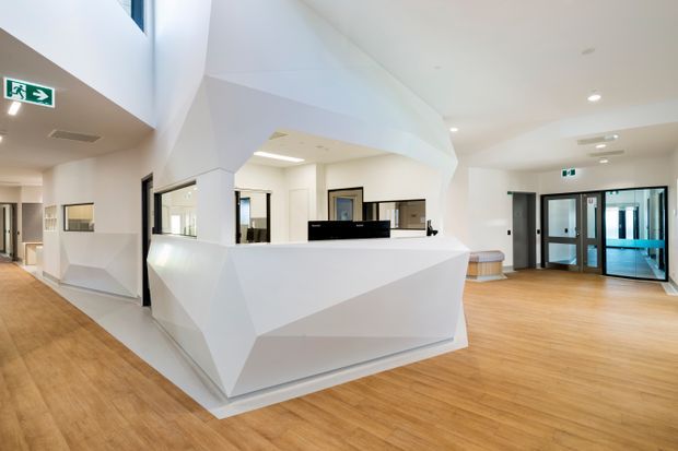 Bright contemporary commercial reception or service area with a sculptural faceted white desk, open pass-through windows, pale timber-look flooring, glass entry doors, and white walls. The open circulation space has recessed lighting and a clean, institutional fit-out.