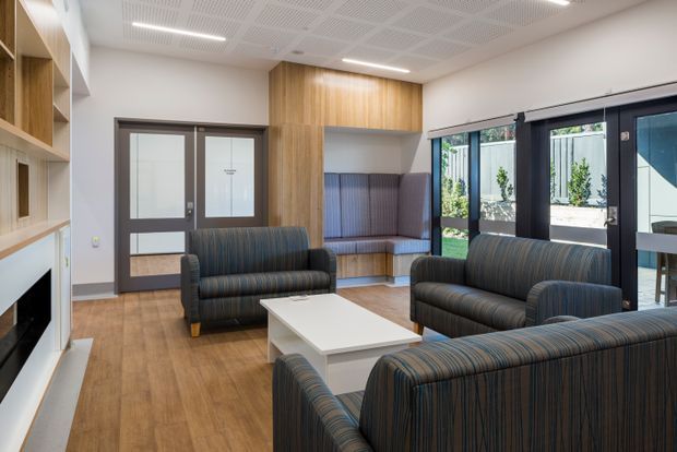 A bright contemporary lounge or waiting area with multiple dark striped upholstered sofas arranged around a white rectangular coffee table. Warm timber flooring, built-in timber shelving and cabinetry, a timber-lined feature wall with integrated bench seating, dark-framed glazed doors and windows, and a white acoustic-panel ceiling define the space. Outdoor greenery is visible through the glazing.