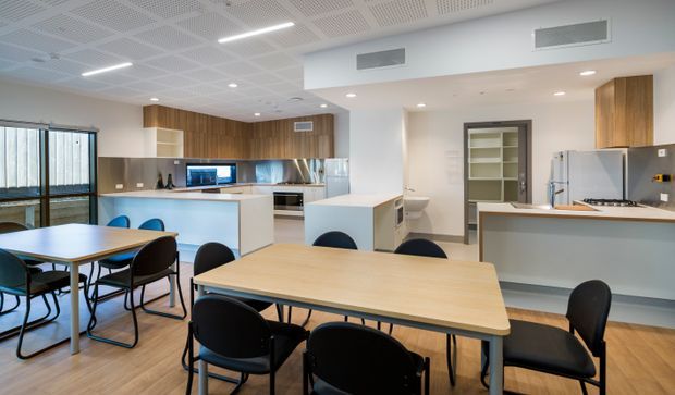 A bright, contemporary communal kitchen and dining area with extensive timber-look cabinetry, pale benchtops, integrated appliances, and multiple large light-wood dining tables surrounded by black chairs. The space has polished timber flooring, white walls, a suspended acoustic ceiling with recessed lighting, large windows and glazed doors, and a central island-style work surface. It appears suited to a shared workplace, education, or institutional setting.