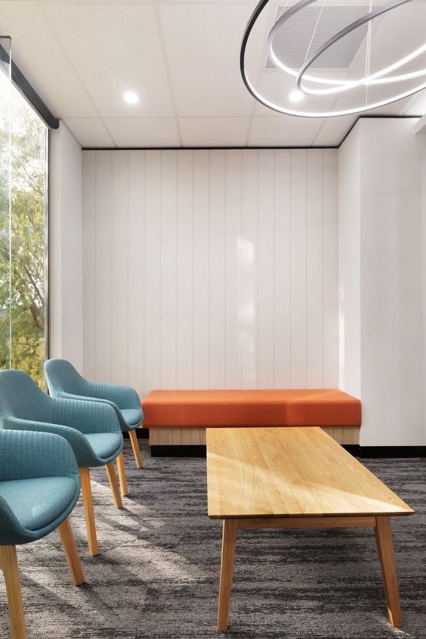 Bright contemporary waiting or lounge area with blue upholstered armchairs, a long orange upholstered bench, and a light timber coffee table. The room features white vertically panelled walls, a suspended sculptural LED ring light, large side glazing, and dark patterned carpet.