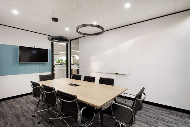 Contemporary commercial meeting room with a large light timber conference table surrounded by black office chairs. The room features a full-width whiteboard, wall-mounted television, blue vertical wall panelling, glass partition doors, circular suspended pendant lights, a white acoustic ceiling, and dark patterned carpet flooring.