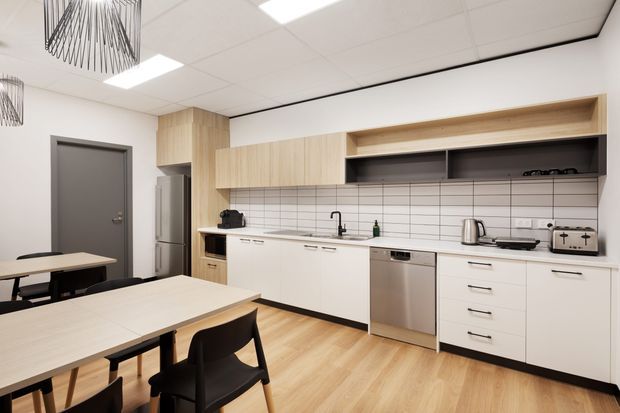 Bright, functional open-plan kitchenette and dining area in a commercial or workplace setting. The space features white flat-panel cabinetry, light timber overhead joinery, a white tiled splashback, stainless-steel appliances, light timber-look flooring, black dining chairs, and contemporary pendant lighting.