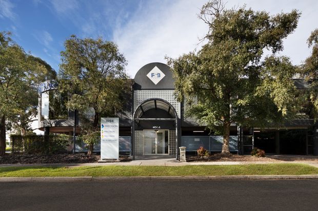 Street-facing view of a low-rise commercial building set among mature trees. The facade combines dark masonry or cladding with a prominent central arched entry canopy, glazed doors, a curved upper feature, and visible business signage.