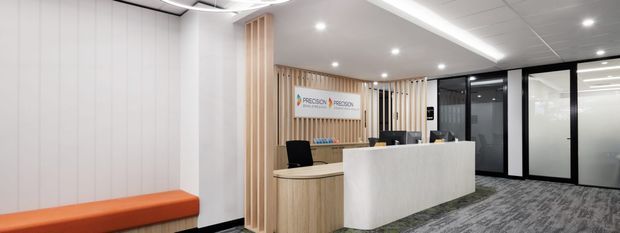 A contemporary commercial reception area with a large white stone-look reception desk, curved light-wood counter, vertical timber slat feature wall, white wall panelling, black-framed glazed office partitions, recessed ceiling lights, and grey patterned carpet. The space has a clean, professional corporate fit-out.