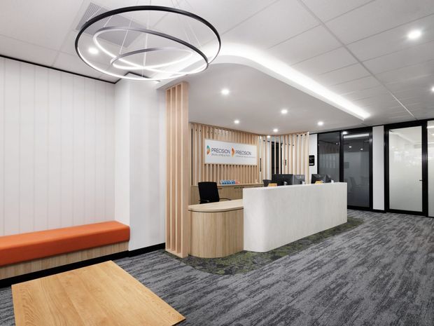 A contemporary commercial reception area with a curved white reception desk, timber-look cabinetry, vertical timber slat feature wall, black-framed glazed doors, patterned grey carpet, and sculptural circular pendant lighting. The space is bright, clean, and professionally fitted out, with orange built-in bench seating and a branded wall behind the desk.