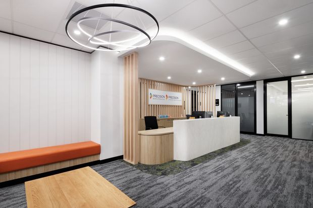 Contemporary commercial reception area with a curved white reception desk, light timber slatted feature wall, integrated orange upholstered bench seating, glass-partitioned offices, patterned carpet, and a large circular sculptural pendant light.