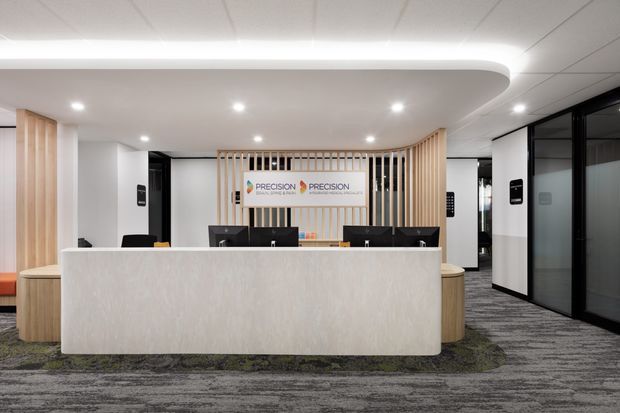A modern commercial reception area with a large curved light-coloured reception desk, black computer monitors, vertical timber slat feature wall, and Precision-branded signage. The space has white walls and ceiling panels, black-framed glazed office partitions, and patterned grey carpet with green accents.