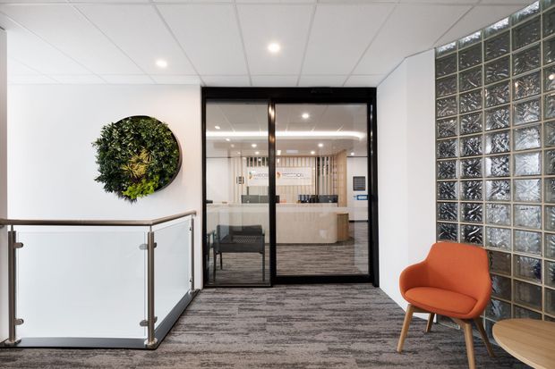 A bright contemporary commercial office reception or landing area with grey carpet, white walls, a tiled suspended ceiling, glass-panel balustrade, black-framed glazed doors, and a textured glass-block feature wall. An orange upholstered lounge chair and round timber table sit beside a circular preserved or artificial greenery wall feature. A reception desk and office area are visible through the glazed doors.