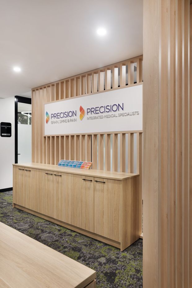 A contemporary medical specialist clinic reception area featuring a light timber reception cabinet and countertop, vertical timber slat screening, integrated illuminated-style branding signage, recessed ceiling lights, and patterned commercial carpet. The space has a clean, professional fit-out with warm timber detailing and a partially visible consultation area beyond.