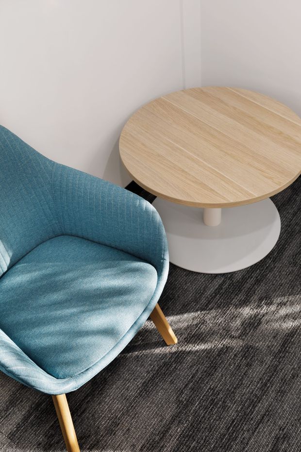 A close-up interior view of a compact seating area featuring a curved blue upholstered lounge chair with light timber legs beside a round light timber coffee table with a white pedestal base. The furnishings sit on a dark textured carpet against plain light walls, with strong natural light and shadows creating a clean, contemporary atmosphere.