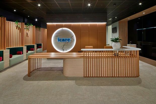A contemporary commercial reception lobby with a prominent timber slatted reception desk, warm wood wall panelling, integrated soft lighting, a circular illuminated logo feature, built-in upholstered seating, and dark ceiling finishes. The space has a refined, warm, hospitality-inspired corporate interior.