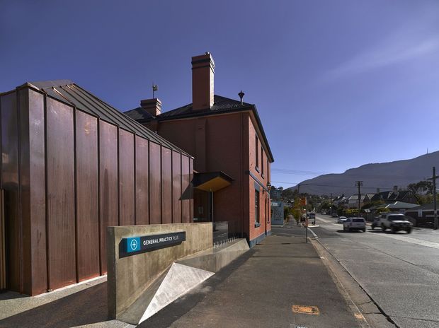 Street-level exterior view of a contemporary medical building integrated with an older red-brick structure. The facade combines tall vertical copper-toned metal panels with exposed brick, a sloped metal roof, and a projecting entry canopy. A sign identifies the premises as a general practice clinic. The building fronts a suburban street with a mountain visible in the distance.