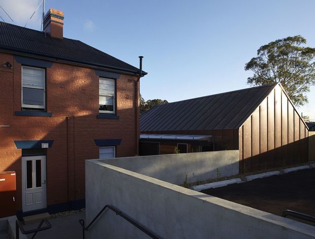 Exterior view of a compact urban architectural project, showing an existing two-storey red-brick building beside a newer dark metal-clad, steeply pitched gable-roofed structure. A rendered concrete boundary wall and stairway occupy the foreground, with mature trees and clear sky beyond.