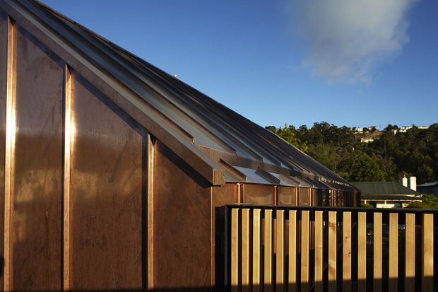 Exterior architectural view of a compact building with a steeply pitched roof and strongly angled roof planes, clad in weathered copper-toned metal with dark metal detailing. A black-framed balcony or deck railing occupies the foreground, with a leafy hillside and neighbouring buildings in the background under a clear sky.