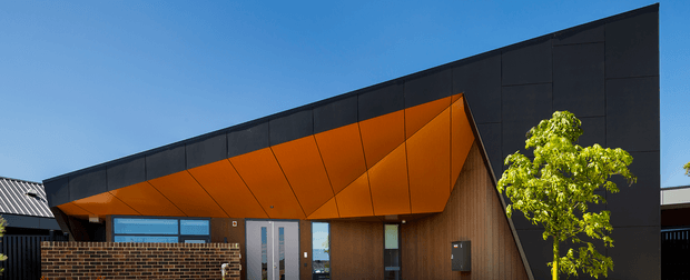 Contemporary single-storey building exterior with a dramatic angular projecting canopy in vivid orange, dark charcoal cladding, warm vertical timber wall panels, and large glazed entry doors. A low brick wall and small landscaped tree sit in front, under a clear blue sky.
