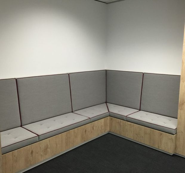 A compact, plain interior room with an L-shaped built-in upholstered bench wrapping around two walls. The seating has light grey fabric cushions and back panels with dark piping, set over timber-look built-in cabinetry. White walls, a dark carpeted floor, and a simple suspended ceiling create a functional, understated workspace or meeting area.