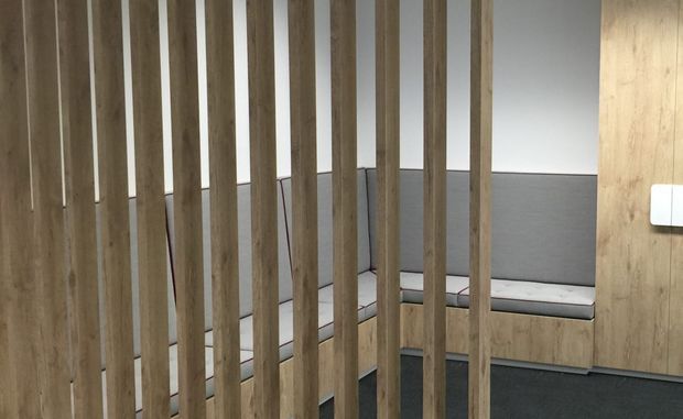 A contemporary interior featuring a vertical timber slat screen in the foreground, partially enclosing a built-in grey upholstered bench around a corner. Timber wall panelling and dark carpet create a warm, understated workspace or communal seating area.